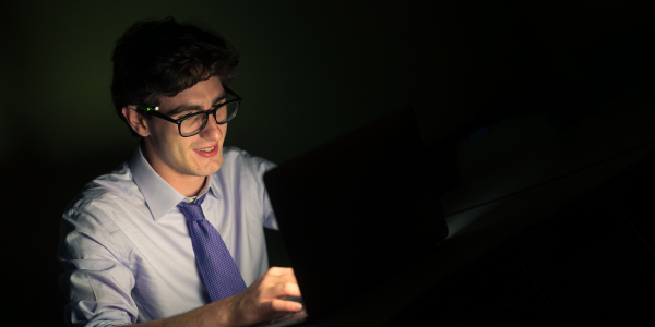 A white man with dark hair and a blue tie reads from a laptop.