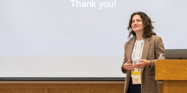 A white woman with brown hair, wearing a tweed blazer, presents at a conference.