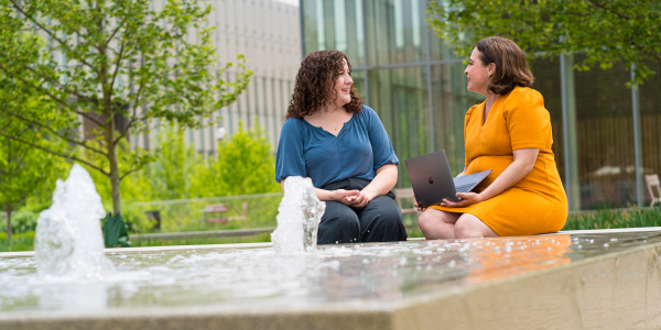 A Latinx professor in a yellow dress with a MacBook, and a white student in a blue shirt discuss graduate education with a fountain in the foreground.