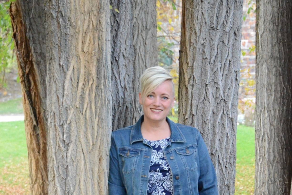 A white woman with short blond hair poses for a photo leaned against a tree.