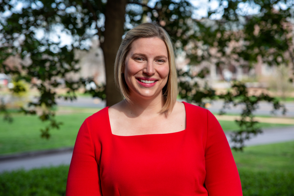 A white woman with a blonde bob is posed for a professional headshot. She is wearing bright red, and stands in from of trees and grass.