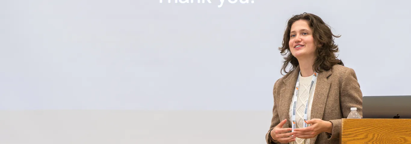 A white woman with brown hair, wearing a tweed blazer, presents at a conference.