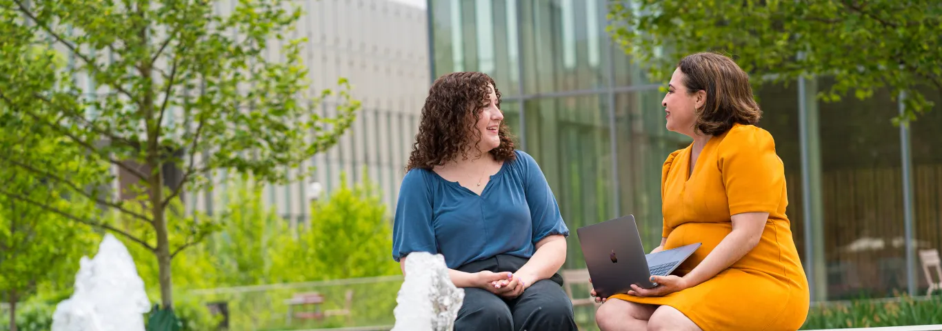 A Latinx professor in a yellow dress with a MacBook, and a white student in a blue shirt discuss graduate education with a fountain in the foreground.