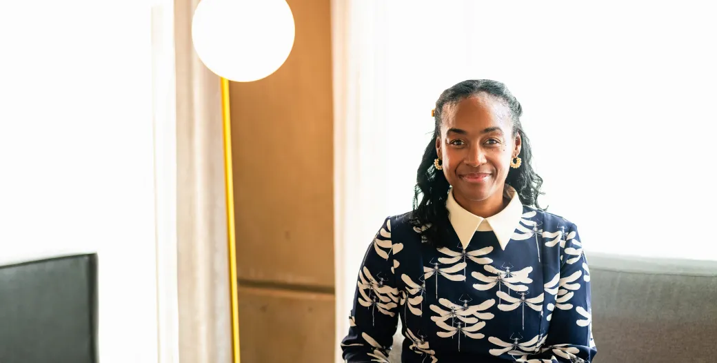 "A Black woman smiles for a professional headshot. She is sitting on a great couch in modern room flooded with daylight. She is wearing a navy blue dress with a dragonfly pattern."