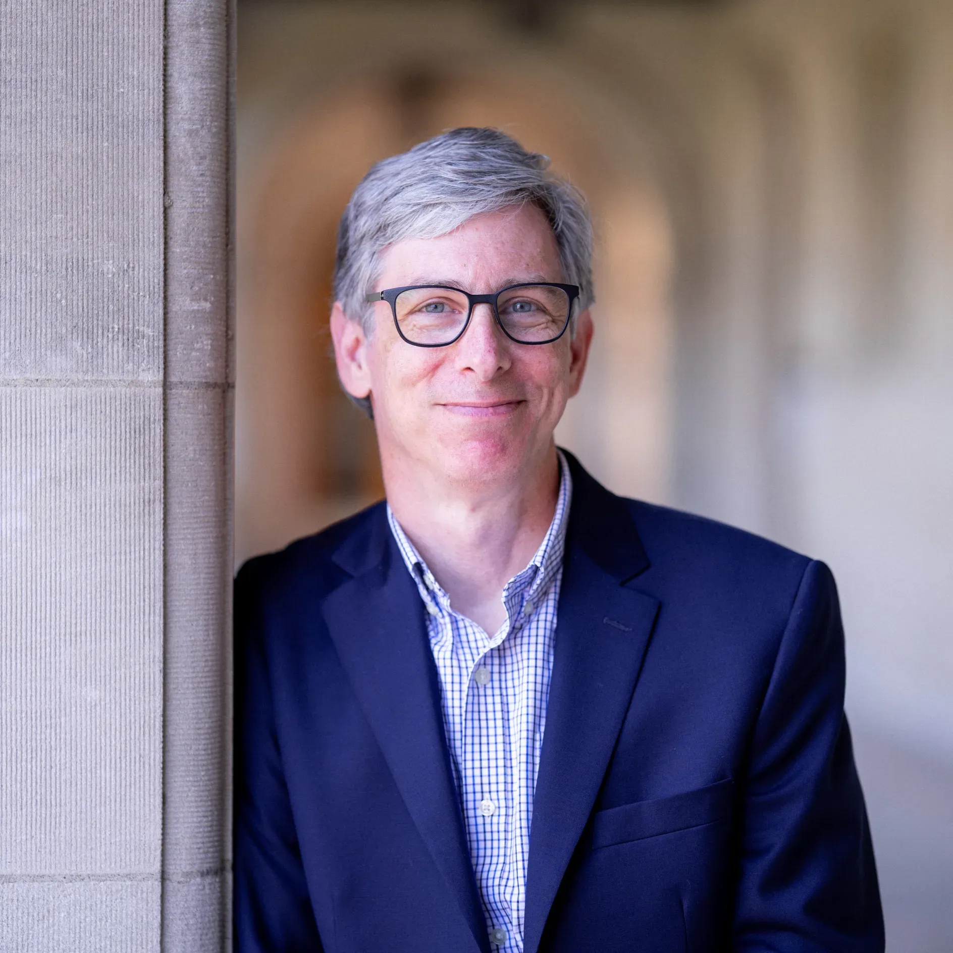 "A white man with grey hair leans on a stone pillar, smiling for a professional headshot. He wears glasses and a deep blue sport coat."