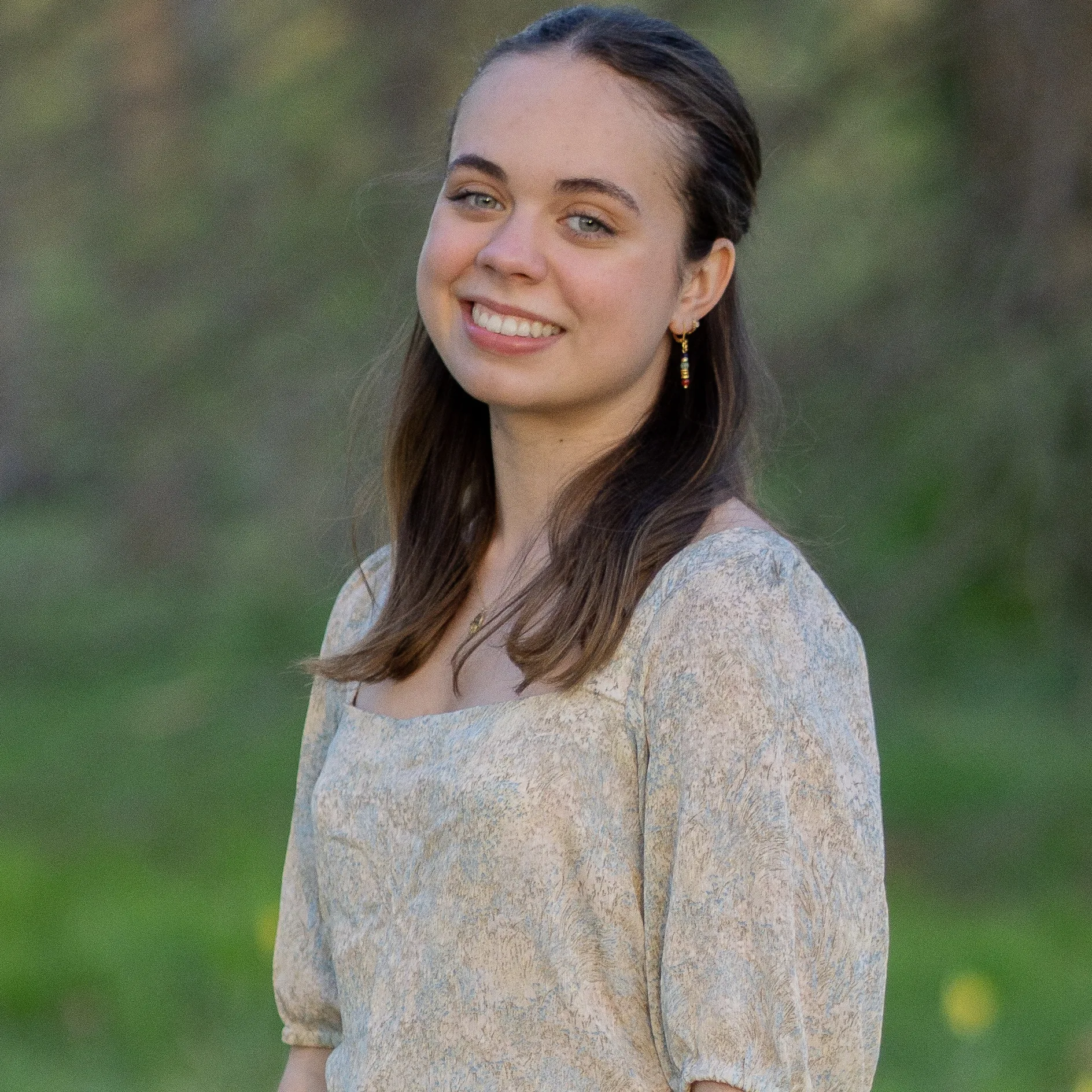 "A white woman with brown hair smiles for a headshot. The background is blurred green vegetation.