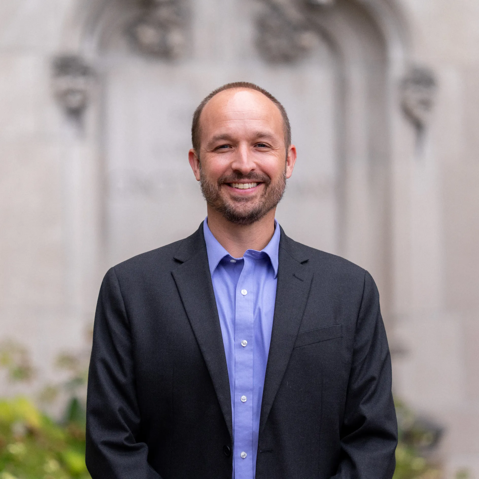 A white man smiles for a professional headshot. He wears an Oxford blue shirt and navy sport coat.