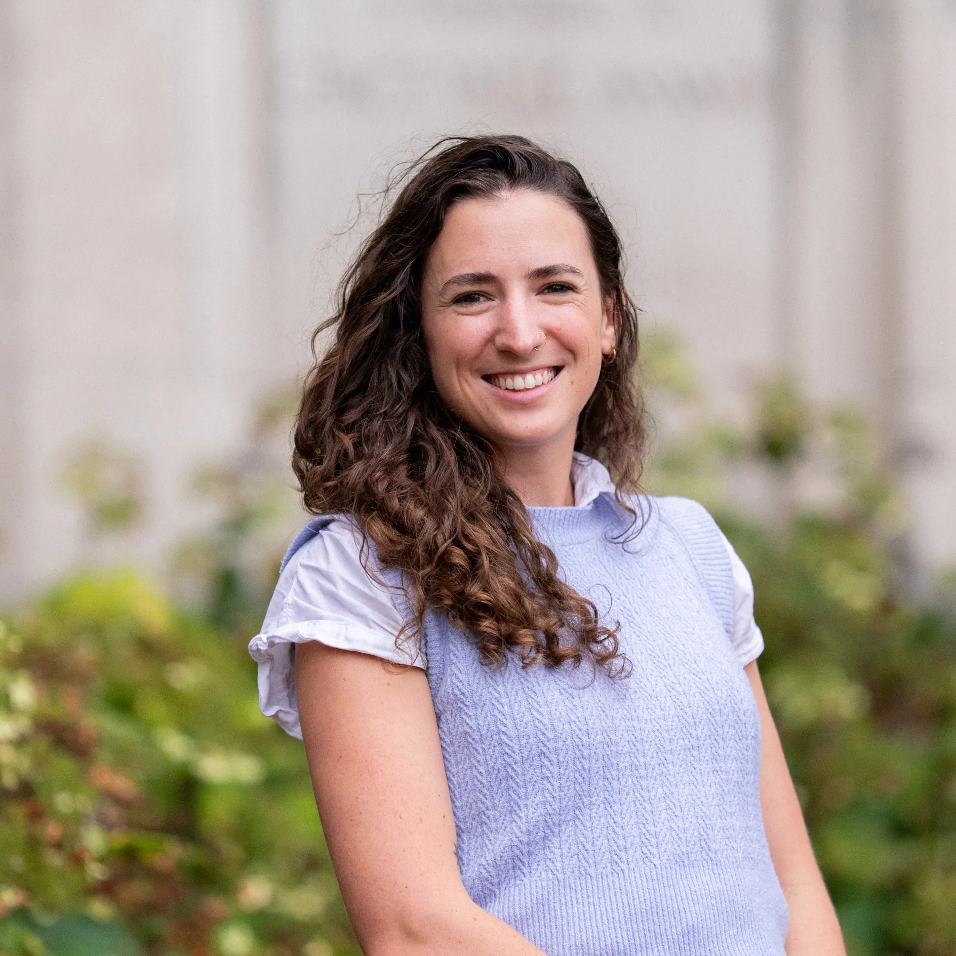 A white woman with brown curly hair poses in front of a beige stone wall and bushes.