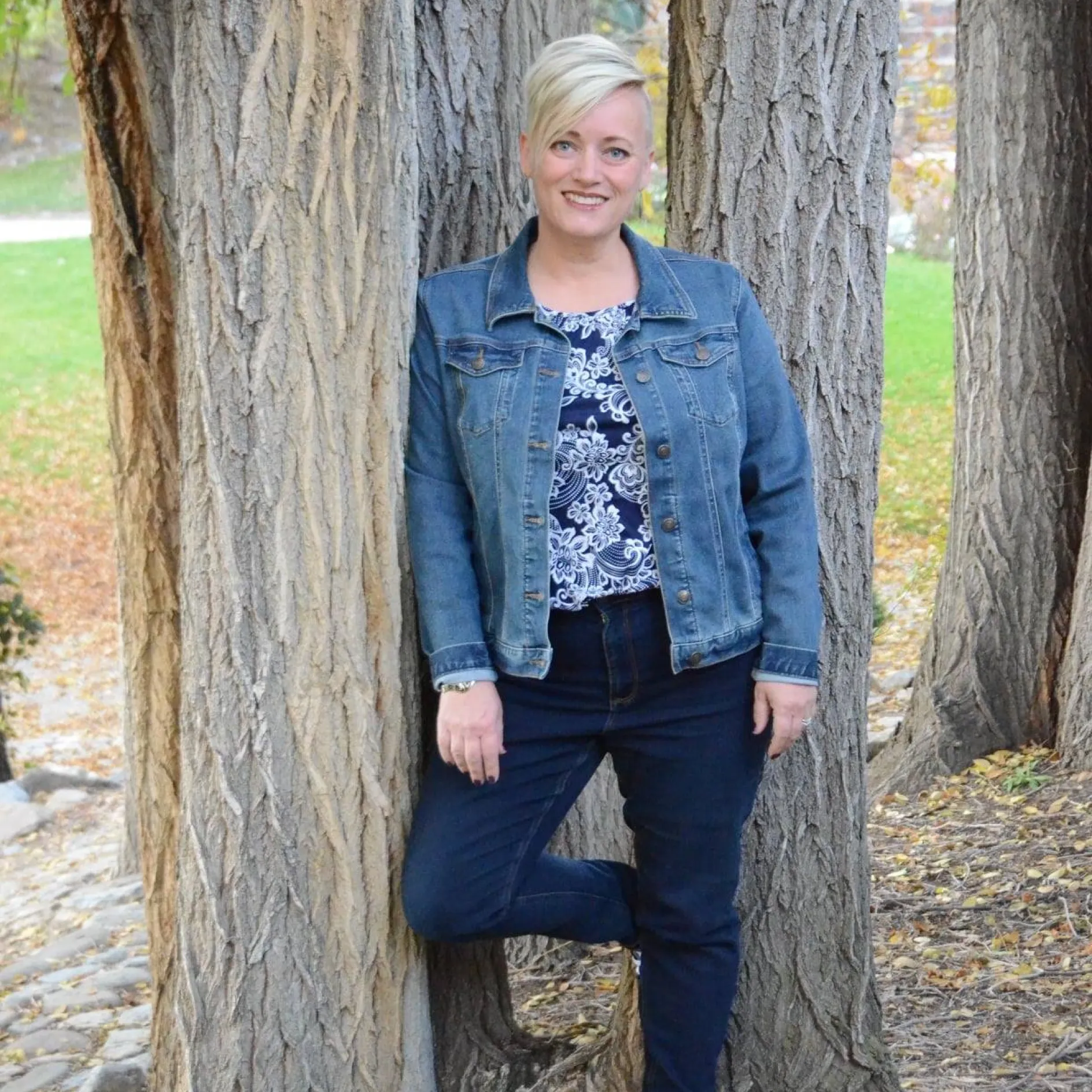 A white woman with short blond hair poses for a photo leaned against a tree.