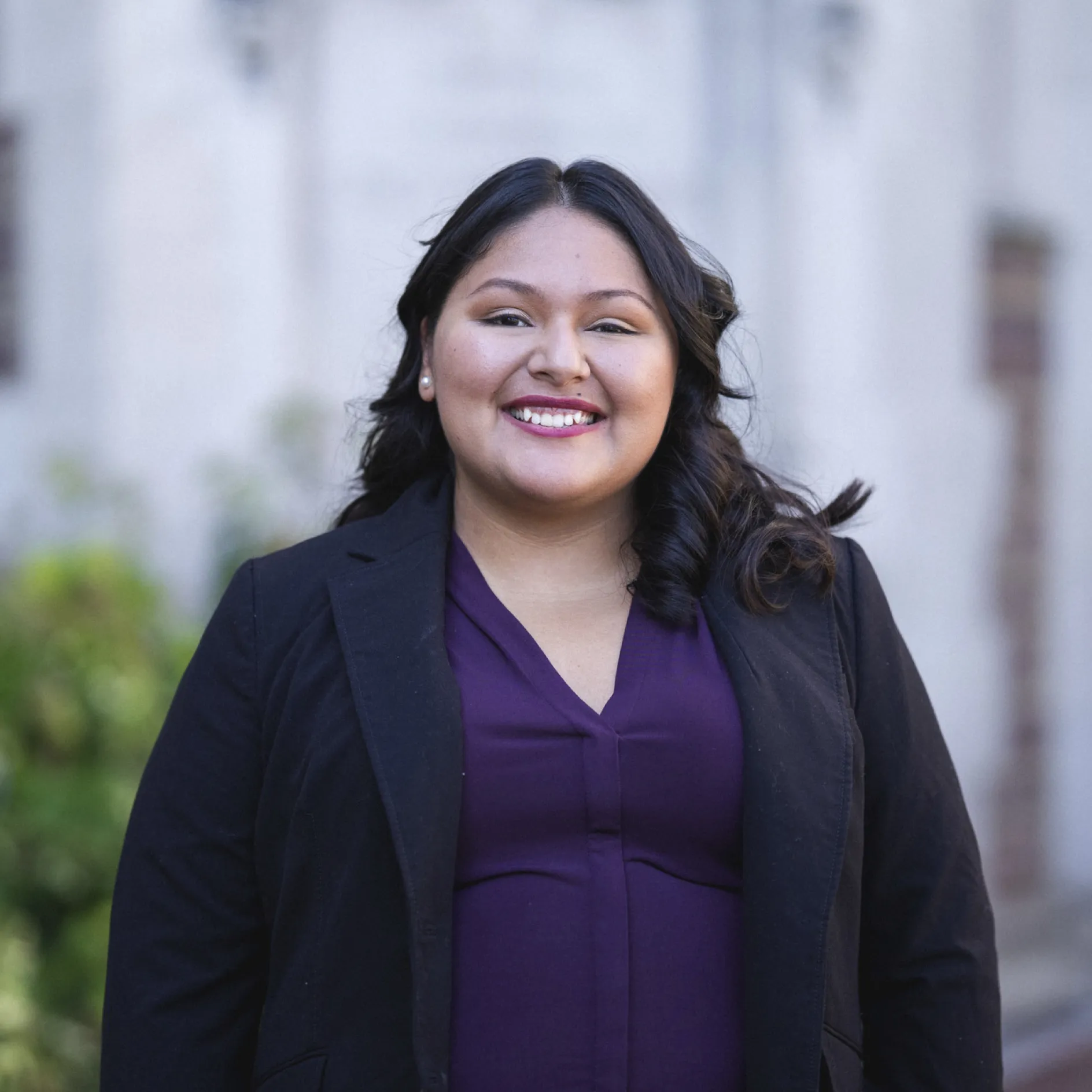 A woman with dark hair and olive skin smiles and poses in front bushes and a stone wall.