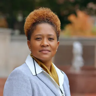 "A Black woman with copper hair is posed for a professional headshot in front of a grass and a brick fountain"