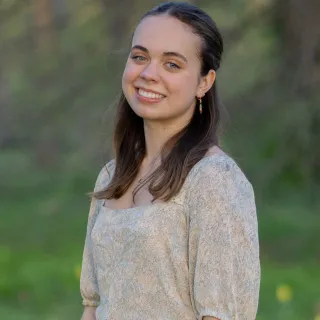 "A white woman with brown hair smiles for a headshot. The background is blurred green vegetation.