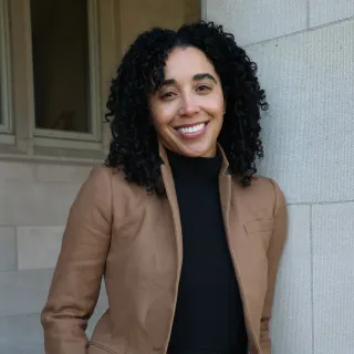 "A woman with light brown skin and black curly hair smiles for a professional headshot. She is wearing a black turtleneck and brown jacket."