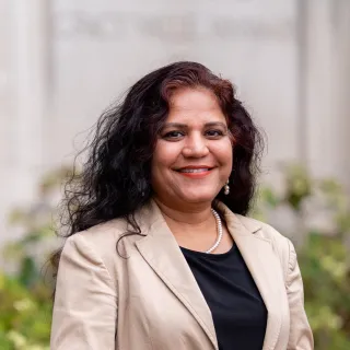A woman with dark curly hair and tan skin poses in front of bushes and a beige stone wall.