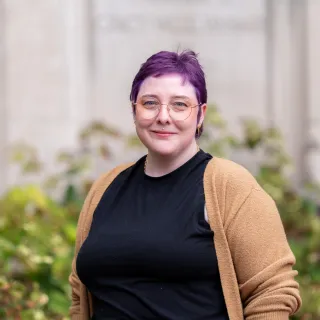 A white, androgenous person with purple hair poses in front of a beige stone wall and green bushes.