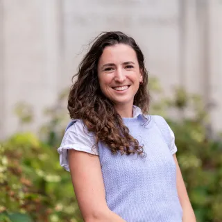 A white woman with brown curly hair poses in front of a beige stone wall and bushes.