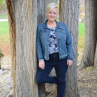 A white woman with short blond hair poses for a photo leaned against a tree.