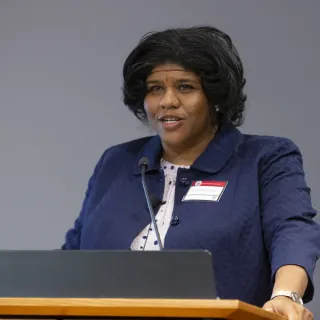 Michelle Purdy, a Black woman with dark hair and a navy blazer, speaks at a podium at the Day of Dialogue and Action
